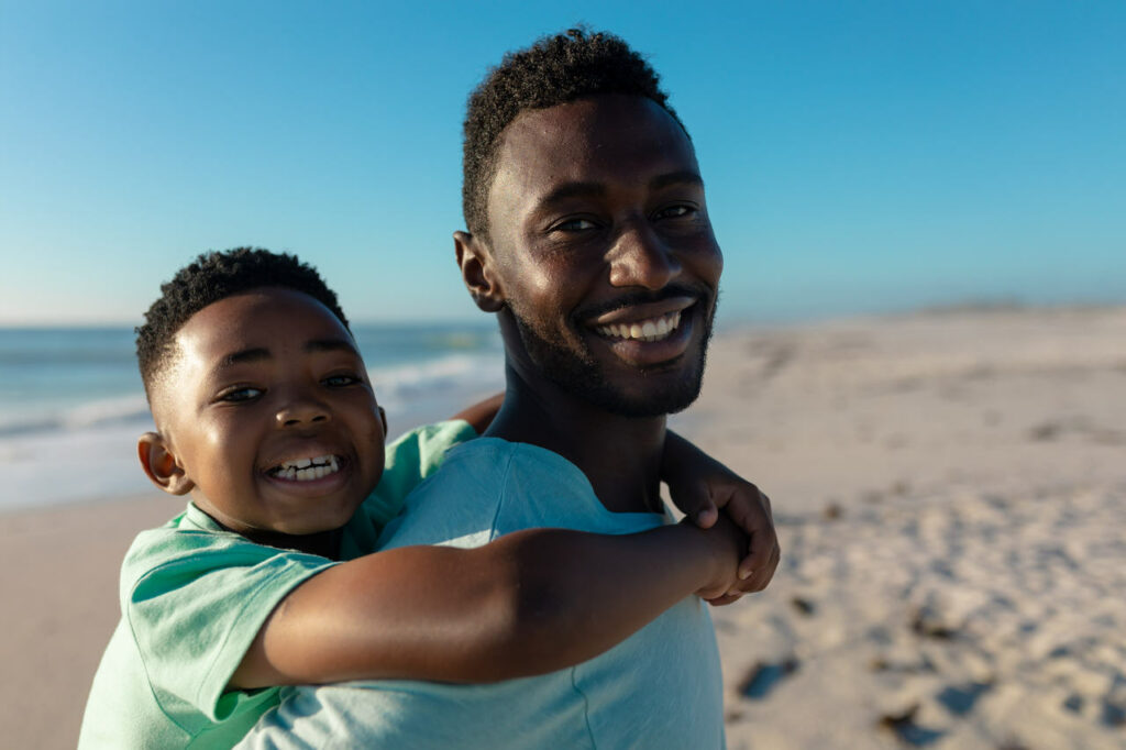 Em uma praia, um homem está carregando um menino nas costas. Ambos estão sorrindo e presume-se que são pai e filho.