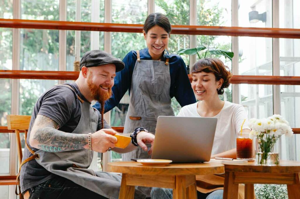 Na imagem, três pessoas - duas mulheres e um homem - conversam e sorriem em frente a um notebook